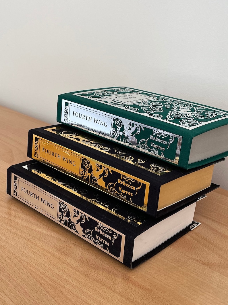Op de afbeelding: Drie hardcover boeken gestapeld op elkaar. Het bovenste boek is groen met zilveren letters en de woorden "Fourth Wing" op de rug. Het middelste boek is zwart met gouden letters en de woorden "Fourth Wing" op de rug. Het onderste boek is zwart met zilveren letters en de woorden "Fourth Wing" op de rug. Alle drie boeken hebben de naam van de auteur, "Rebecca Yarros", op de rug gedrukt.