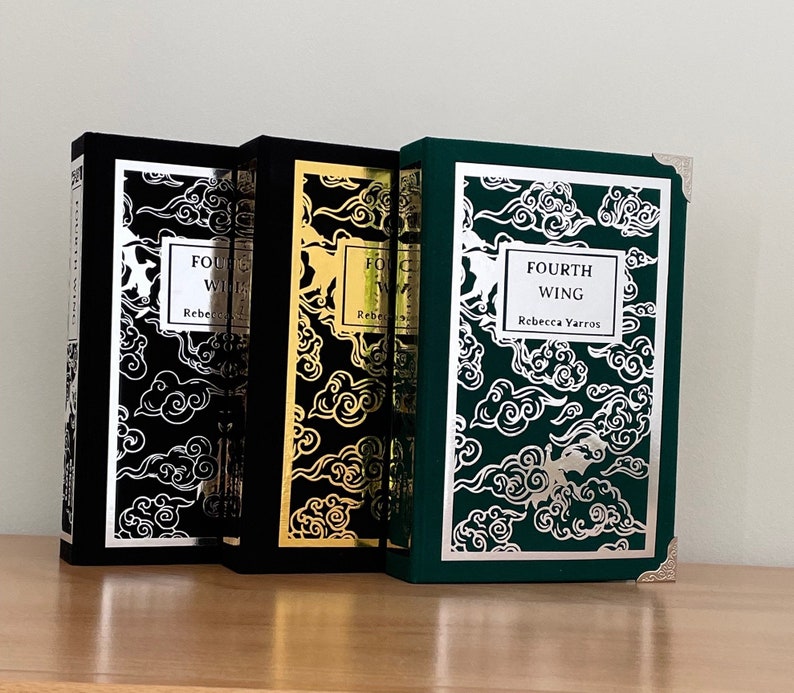 Op de afbeelding: Drie hardcover boeken met zwarte, gouden en groene omslagen. De omslagen hebben een zilveren wolkenpatroon en de tekst "Fourth Wing" van Rebecca Yarros.