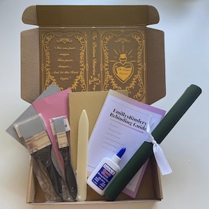Pu&ograve; includere: Una scatola di cartone marrone piena di forniture per la rilegatura. La scatola contiene due pennelli, un osso per piegare, una bottiglia di colla, due pezzi di carta vetrata, un pezzo di carta rosa, un pezzo di carta marrone, un tessuto verde, un nastro bianco e una guida intitolata "Emillys Bindery Rebinding Guide".