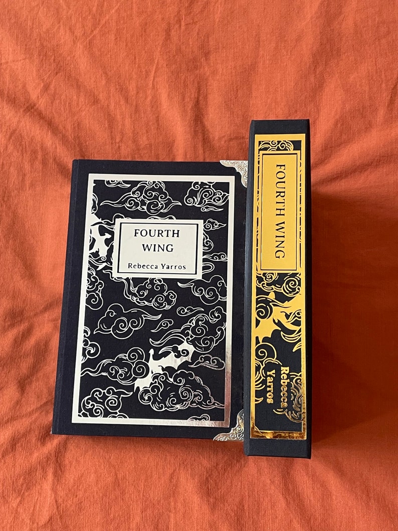 Op de afbeelding: Een zwart hardback boek met een zilveren en zwarte omslag met een wolkendesign. De titel van het boek is "FOURTH WING" en de auteur is "Rebecca Yarros". Het boek ligt naast een zwarte rug met gouden letters die "FOURTH WING" en "Rebecca Yarros" lezen.
