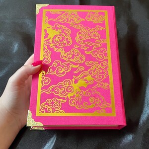 Op de afbeelding: Een roze hardcover boek met gouden decoratieve hoeken en een gouden wolkendesign op de omslag.