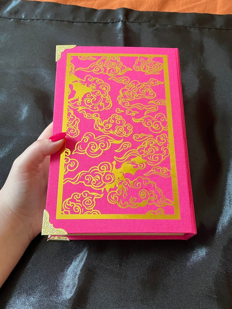 Op de afbeelding: Een roze hardcover boek met gouden decoratieve hoeken en een gouden wolkendesign op de omslag.