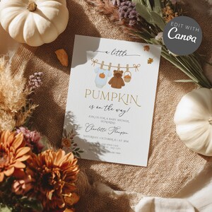 Op de afbeelding: Een uitnodiging voor een babyshower met de tekst "Pumpkin is on the way!" in gouden letters. De uitnodiging bevat illustraties van babykleding, een teddybeer en een mand. Het ontwerp bevat bloemaccenten en een witte pompoen.