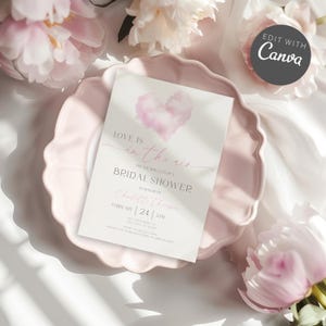 Può includere: Un invito per l'addio al nubilato con la scritta "Love is in the air" e un cuore ad acquerello. L'invito è su un piatto rosa, circondato da fiori rosa. L'invito include la data e l'ora.