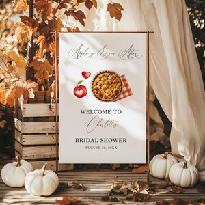 Op de afbeelding: Een welkomstbord voor een bruidsdouche met de tekst "Apple-y Ever After". Het bord toont illustraties van een appeltaart, een rode appel en een hart. Het bord staat op een koperkleurig metalen frame, met witte pompoenen en herfstbladeren op de voorgrond.