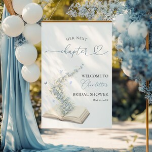 May include: A welcome sign for a bridal shower, with the text "Her Next Chapter" and "Welcome to Charlotte's Bridal Shower". The sign has a floral design with an open book and butterflies, with a gold frame. White balloons and blue fabric are in the background.