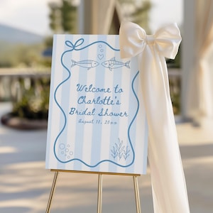 Pode incluir: Uma placa de boas-vindas para um chá de panela, com um fundo listrado azul claro e branco. A placa exibe o texto "Welcome to Charlotte's Bridal Shower" e a data. Uma fita creme adorna a lateral. A placa está em um cavalete dourado.