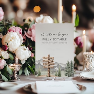 Op de afbeelding: Een aangepast bord met de tekst "Custom Sign" en "FULLY EDITABLE". Het bord toont een aquarel illustratie van een houten wegwijzer met pijlen die wijzen naar "Love Ahead", "Prosecco Peak", "I Do Summit" en "Bridal Basecamp".