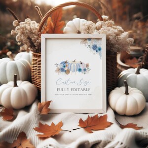 Op de afbeelding: Een ingelijst bord met de woorden "Custom Sign" en "Fully Editable" is omgeven door witte pompoenen, herfstbladeren en een rieten mand. Het bord heeft een aquarelontwerp van pompoenen en blauwe bloemen.