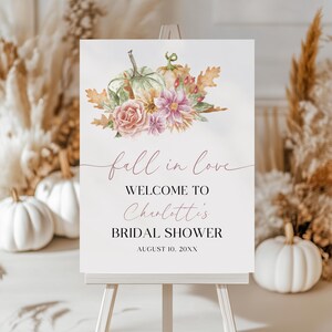 Op de afbeelding: Een wit bord op een ezel met de woorden "fall in love" en "Welcome to Charlotte's Bridal Shower" in een scriptlettertype. Het bord is versierd met aquarel pompoenen, bloemen en bladeren.