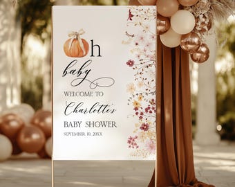 May include: A welcome sign for a baby shower with a pumpkin illustration and floral accents. The sign reads "Oh baby, Welcome to Charlotte's Baby Shower, September 10, 20XX". The sign is on a gold metal stand, with balloons and fabric in the background.