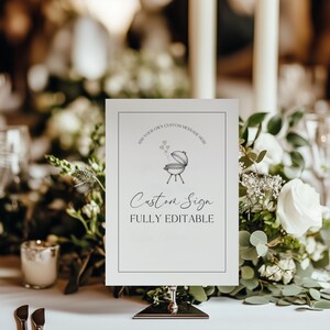 Op de afbeelding: Een wit bord met een zwarte rand, met een barbecue-illustratie en de tekst "Custom Sign - Fully Editable". Het bord wordt gepresenteerd op een metalen standaard, omringd door bloemstukken, ideaal voor evenementen.