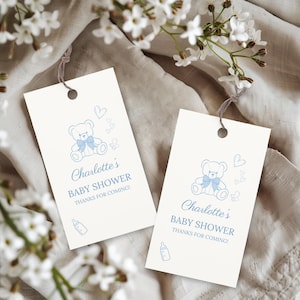 Op de afbeelding: Twee crèmekleurige baby shower cadeaulabels met een blauwe teddybeer illustratie, hartjes en een babyfles. De tekst luidt "Charlotte's BABY SHOWER THANKS FOR COMING!". Elk label heeft een gat aan de bovenkant met een bruin touwtje.