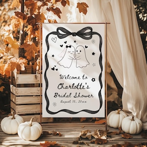 Op de afbeelding: Een wit bord met een zwarte lintrand en strik, dat gasten verwelkomt op Charlotte's Bridal Shower. Het bord toont twee cartoongeesten, een met een sluier, en decoratieve harten, sterren en bloemen. Het bord staat op een koperkleurige standaard.