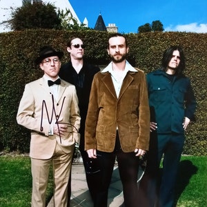 Puede incluir: Cuatro hombres de pie frente a un seto, uno lleva una chaqueta de traje marrón, otro lleva un traje beige, uno lleva una camisa azul y otro lleva una camisa negra. El hombre con el traje beige sostiene un micrófono.