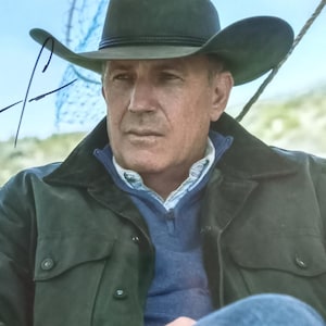 Könnte beinhalten: Porträt eines Mannes mit einem dunkelgrünen Cowboyhut und einer Jacke über einem blauen Pullover. Das Bild enthält ein Autogramm in der oberen linken Ecke. Der Mann schaut zur Seite.