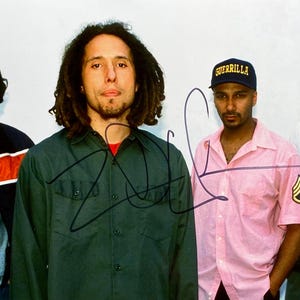May include: A group of four people posing for a photograph. One person wears a dark green button-up shirt, and another wears a pink short-sleeved shirt with a patch on the sleeve and a navy blue cap with the word "GUERRILLA". The image is signed.