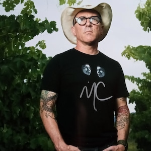 Puede incluir: Un hombre con una camiseta negra con un gráfico de dos caras y la firma "i/c". Lleva un sombrero de vaquero blanco y gafas. El hombre está de pie frente a un fondo verde frondoso.