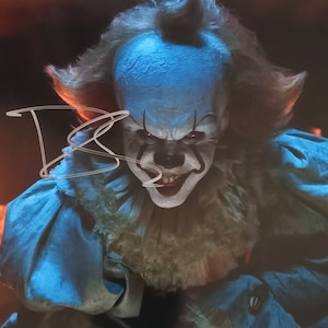 May include: A close-up of a menacing clown with white face paint, red accents, and a sinister grin. The clown has blue hair and a ruffled collar. The image includes a signature in the upper left corner.