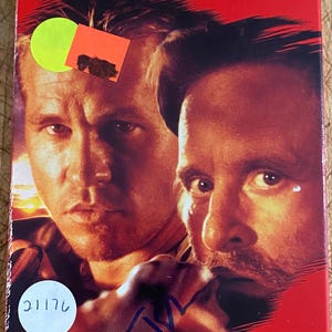 Puede incluir: Una portada de VHS con Michael Douglas y Val Kilmer en un primer plano. El título de la película es "The Ghost and the Darkness" con el lema "Prey for the Hunters".