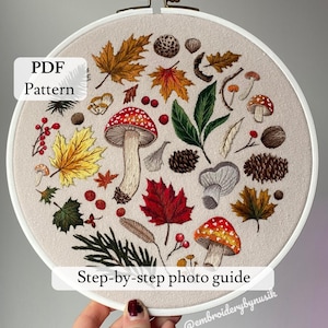 May include: Embroidered hoop art featuring a variety of autumnal elements, including mushrooms, leaves, berries, and pine cones. The hoop is white with a beige fabric background. The text "PDF Pattern" and "Step-by-step photo guide" are visible on the image. The text "@embroiderybynusik" is also visible on the image.