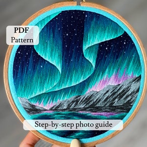 May include: Embroidered hoop art featuring a colorful aurora borealis scene with a dark blue sky, green and purple lights, and a dark gray mountain range reflected in a still body of water. The text "PDF Pattern" and "Step-by-step photo guide" are visible below the image.