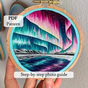 May include: A close-up of a finished embroidery hoop with a colorful depiction of the aurora borealis over a lake. The hoop is made of wood and has a blue felt backing. The text "PDF Pattern" and "Step-by-step photo guide" are visible below the hoop.