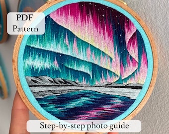 May include: A close-up of a finished embroidery hoop with a colorful depiction of the aurora borealis over a lake. The hoop is made of wood and has a blue felt backing. The text "PDF Pattern" and "Step-by-step photo guide" are visible below the hoop.