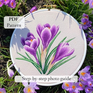 May include: An embroidery hoop with a floral design featuring purple crocus flowers, green leaves, and tan stems. The text "PDF Pattern" and "Step-by-step photo guide" are visible. The hoop is surrounded by crocus flowers.