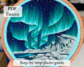 May include: A close-up of a finished embroidery hoop with a design of the aurora borealis over a snowy mountain range. The hoop is made of wood and has a light blue fabric backing. The text "PDF Pattern" and "Step-by-step photo guide" are visible on the hoop.