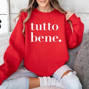 Pode incluir: Um moletom vermelho com as palavras "tutto bene." em branco, centralizadas na frente. O moletom é de gola redonda. A pessoa na imagem está usando jeans claros com joelhos rasgados.