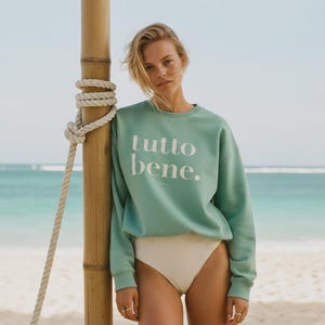 Könnte beinhalten: Hellblaues Sweatshirt mit dem weißen Schriftzug „tutto bene.“. Das Sweatshirt wird mit einem weißen Badeanzug getragen. Der Hintergrund zeigt eine Strandszene mit einer Bambusstange und einem Seil.
