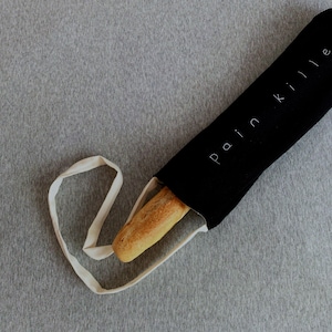 May include: A black baguette bag with the words "pain killer" in white script. A partially visible golden-brown baguette is inside the bag. The bag has a white strap and is set against a gray background.