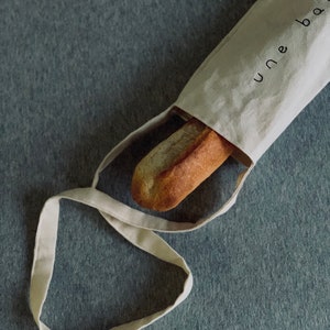 Puede incluir: Una bolsa de tela blanca con el texto "une bag" impreso en ella. Una barra de pan está dentro de la bolsa.