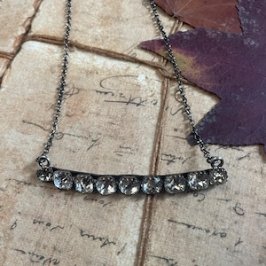Può includere: Una collana color argento con una fila di cristalli trasparenti e scintillanti incastonati in una barra curva. La collana ha una catena delicata ed è esposta su uno sfondo di carta vintage con foglie autunnali.