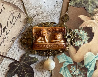 May include: A gold necklace with a cameo featuring two cherubs. The cameo is set in a filigree frame and hangs from a chain with decorative links. A white pearl dangles from the bottom of the necklace.