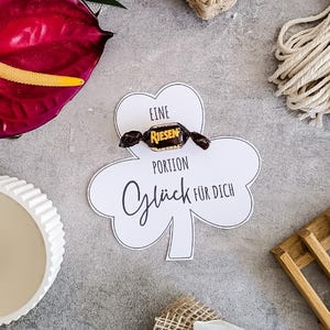 Peut inclure: Une carte blanche en forme de trèfle avec le texte "Eine Portion Glück für Dich" et un bonbon emballé. Une fleur d'anthurium rouge foncé, une bougie blanche et une bobine de corde sont également visibles.