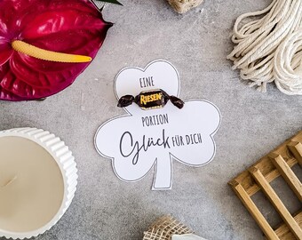 May include: A white clover-shaped card with the text "Eine Portion Glück für Dich" and a wrapped candy. A dark red anthurium flower, a white candle, and a coil of rope are also visible.