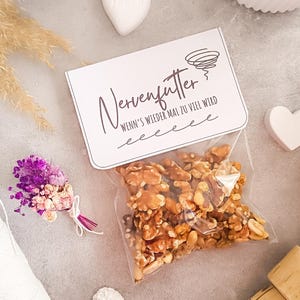 Könnte beinhalten: Ein klarer Cellophanbeutel gefüllt mit Walnüssen und Erdnüssen, mit einer weißen Karte mit der Aufschrift "Nervenfutter" in schwarzer Schrift. Ein kleiner Strauß aus lila und beigen Blumen und eine herzförmige Dekoration sind ebenfalls zu sehen.