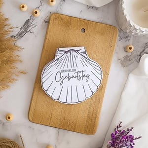 Puede incluir: Una tarjeta blanca en forma de concha con el texto "EINLADUNG ZUM GEBURTSTAG" se coloca sobre una tabla de cortar de madera. La tabla es de color marrón claro con un agujero en la parte superior. El fondo incluye una superficie de mármol, un candelabro blanco y elementos decorativos.