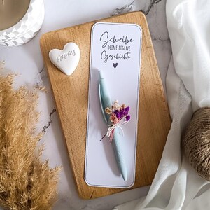 Puede incluir: Un bolígrafo azul claro con un arreglo floral, sobre una tarjeta blanca con el texto "Schreibe deine eigene Geschichte" y un símbolo de corazón. El bolígrafo está sobre una tabla de madera, con un objeto blanco en forma de corazón con "Happy".