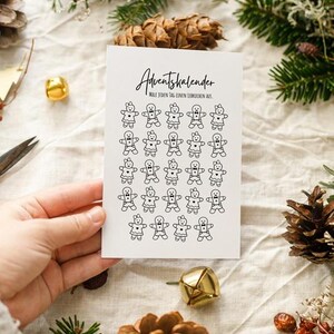Puede incluir: Una tarjeta blanca con el texto alemán "Adventskalender" e ilustraciones de hombrecitos de jengibre. La tarjeta es sostenida por una mano, con decoraciones festivas en el fondo.