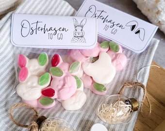 May include: A clear plastic bag filled with pink and white bunny-shaped gummies. The bag has a white label with black text that reads "Osterhasen to go".