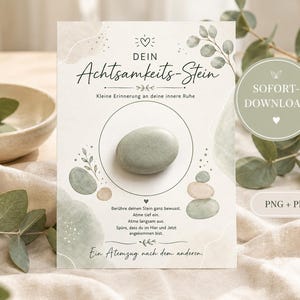 Könnte beinhalten: Eine rechteckige Karte mit dem Text "DEIN Achtsamkeits-Stein" und "Kleine Erinnerung an deine innere Ruhe", die einen glatten, hellgrünen ovalen Stein in einem Kreis zeigt, umgeben von Aquarellillustrationen aus Blättern und Steinen.