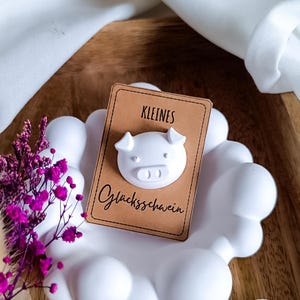 Puede incluir: Un adorno de cerdo de cerámica blanca sobre una tarjeta marrón con las palabras alemanas "KLEINES" y "Glücksschwein" en escritura negra. El adorno está colocado sobre un soporte blanco en forma de nube, con flores moradas en el fondo.