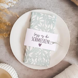 May include: A white plate holds a folded napkin with a leaf pattern and a decorative tag. The tag reads "lass es dir schmecken" with a purple heart. A silver ribbon secures the tag.