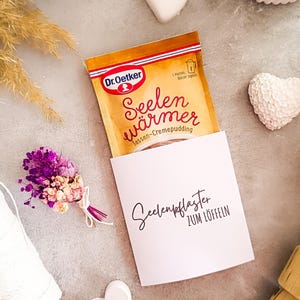Könnte beinhalten: Ein Päckchen Dr. Oetker "Seelenwärmer" Cremepudding in einer weißen Karte mit dem Text "Seelenpflaster zum Löffeln". Das Bild enthält auch einen kleinen Strauß lila und rosa Blumen, ein weißes Herz und dekorative Elemente.