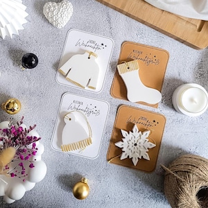 Könnte beinhalten: Weiße Weihnachtsschmuckstücke in Form eines Pullovers, eines Strumpfes, einer Mütze und eines Schneeflocks, jeweils mit goldenen Akzenten. Die Ornamente befinden sich auf Karten mit deutschem Text. Weitere Dekorationen sind eine Kerze, goldene Kugeln und eine Schnurkugel.