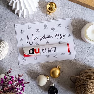 Può includere: Un biglietto di auguri con il testo "Wie schön, dass du DA BIST!" e illustrazioni festive. Una barretta di cioccolato è posta sulla carta. La scena è decorata con decorazioni natalizie, un albero di carta bianco e una candela.