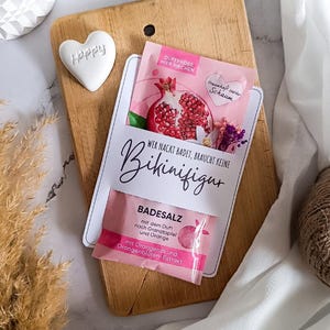 May include: A wooden cutting board with a white heart-shaped decoration that says "Happy". A pink packet of bath salts with the text "WER NACKT BADET, BRAUCHT KEINE BIKINIFIGUR" is displayed. The packet features a pomegranate illustration.